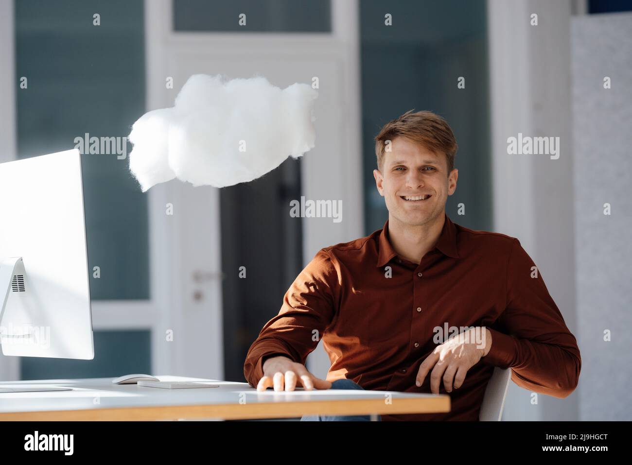 Un uomo d'affari sorridente con un PC desktop che si siede a un livello di rete cloud in ufficio Foto Stock