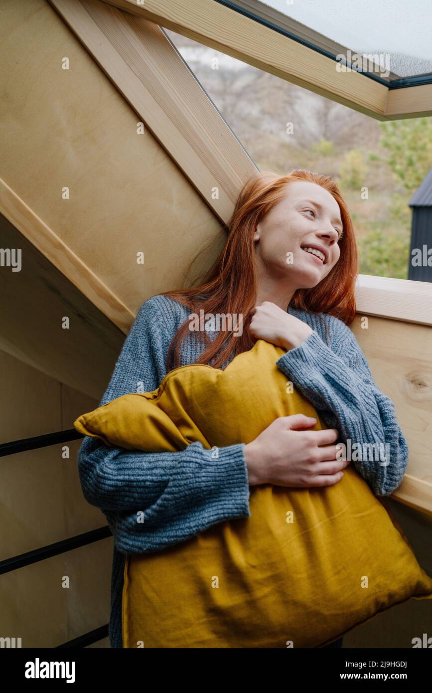 Giovane donna sorridente con cuscino giallo che guarda attraverso la finestra Foto Stock