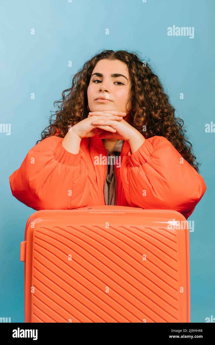 Giovane donna con le mani aggrappate appoggiate sulla valigia arancione su sfondo blu Foto Stock