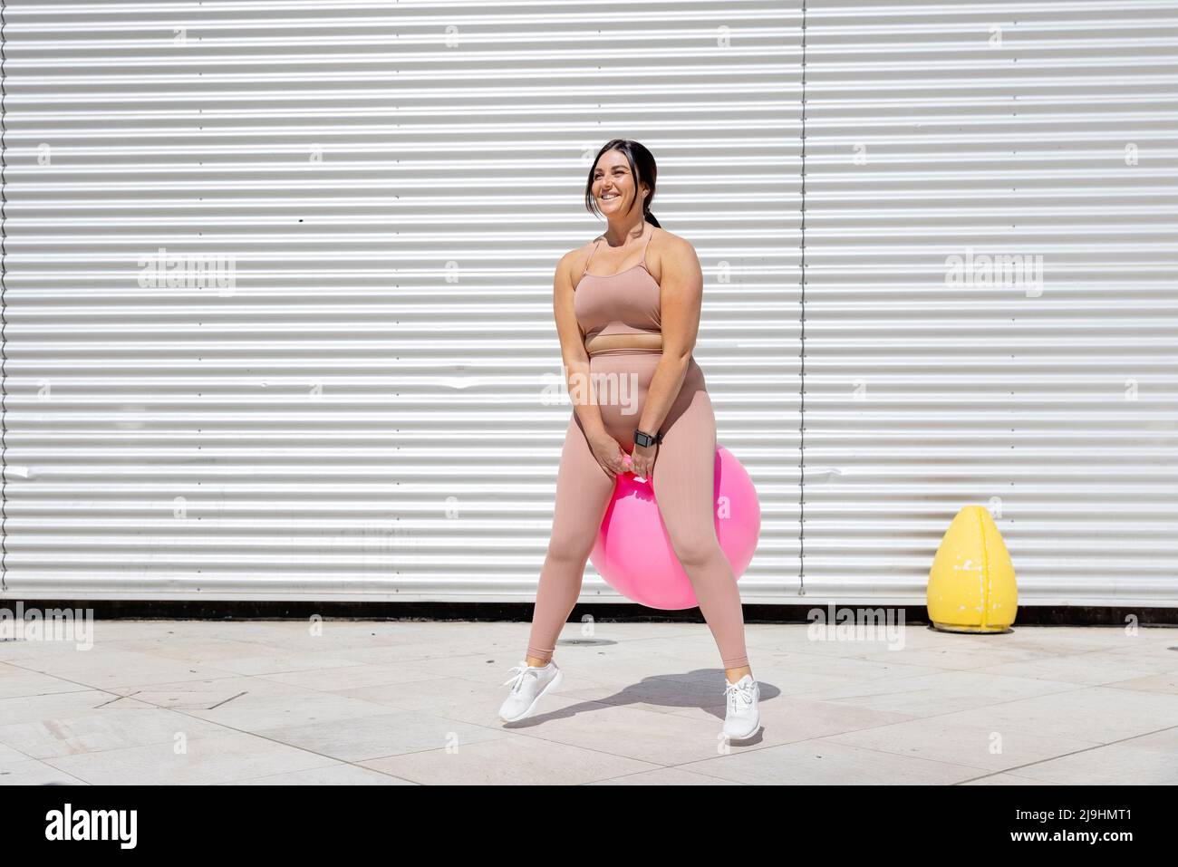 Donna felice curvy esercitandosi con la sfera di idoneità davanti al muro corrugato Foto Stock