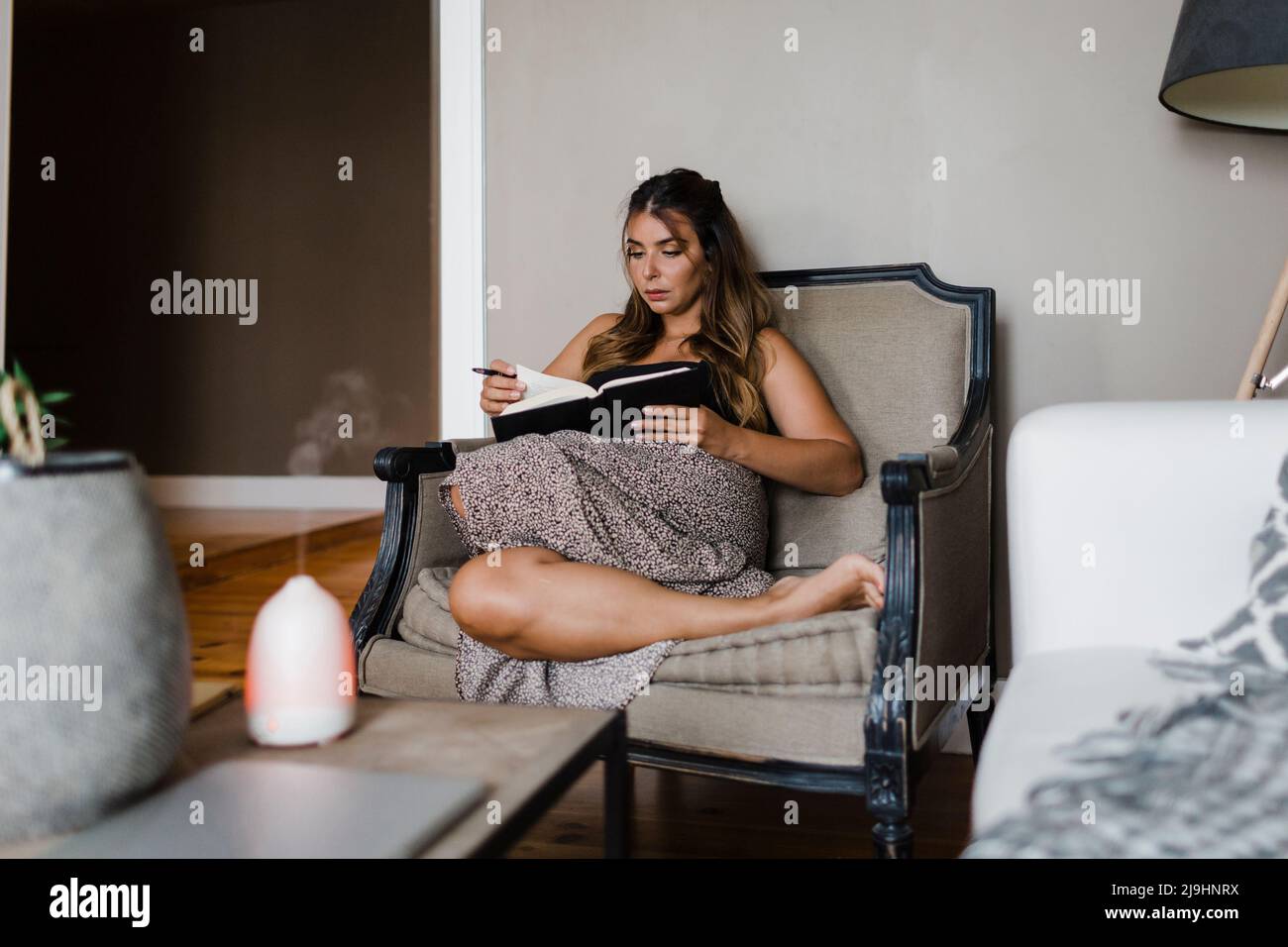 Giovane donna che legge libro seduta in poltrona a casa Foto Stock