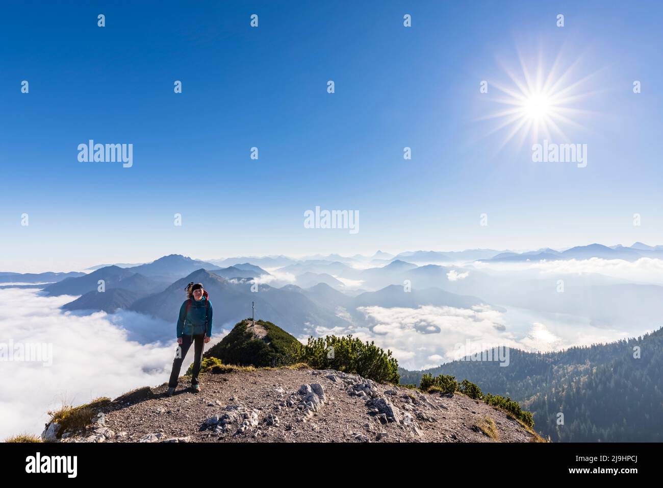 Germania, Baviera, sole che brilla su una donna escursionista in piedi sulla cima della montagna con la nebbia fitta e la vetta del monte Herzogstand sullo sfondo Foto Stock