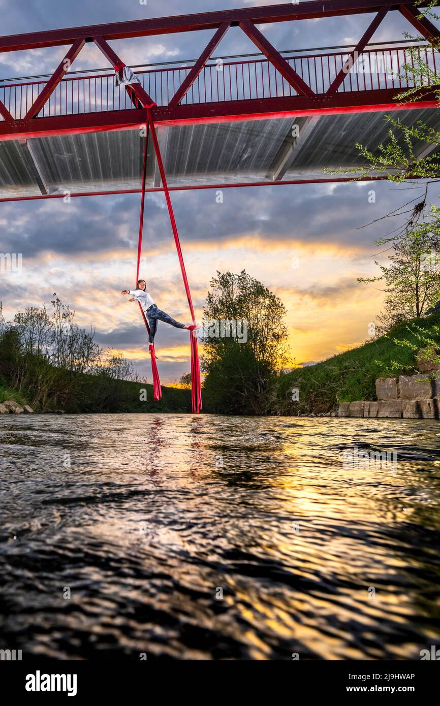 Atleta che esercita acrobatica aerea appeso dal ponte sul fiume Rems al tramonto Foto Stock