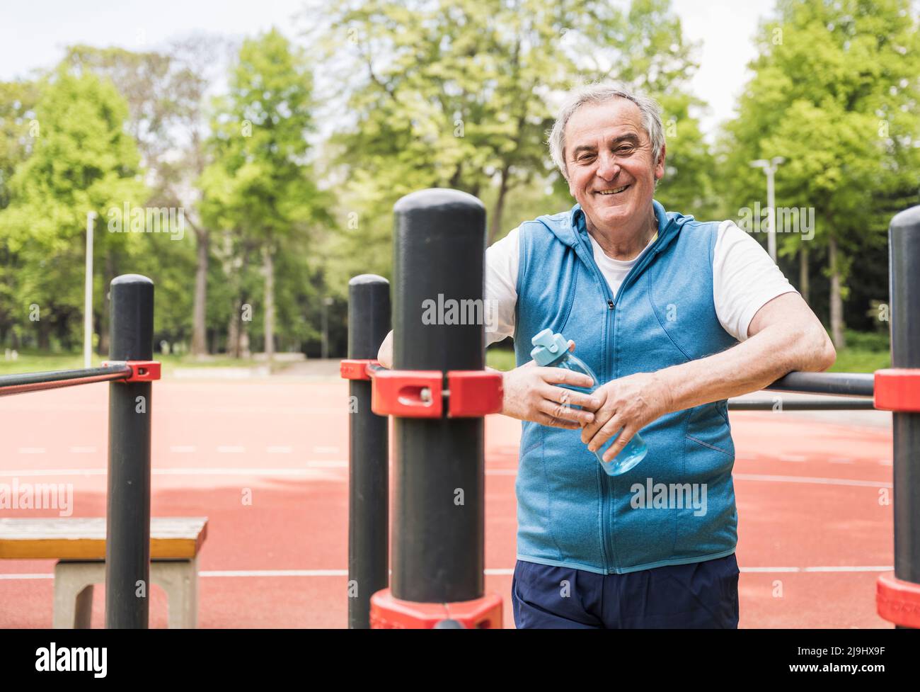 Uomo anziano sorridente che tiene la bottiglia d'acqua in piedi in mezzo al bar di ginnastica Foto Stock