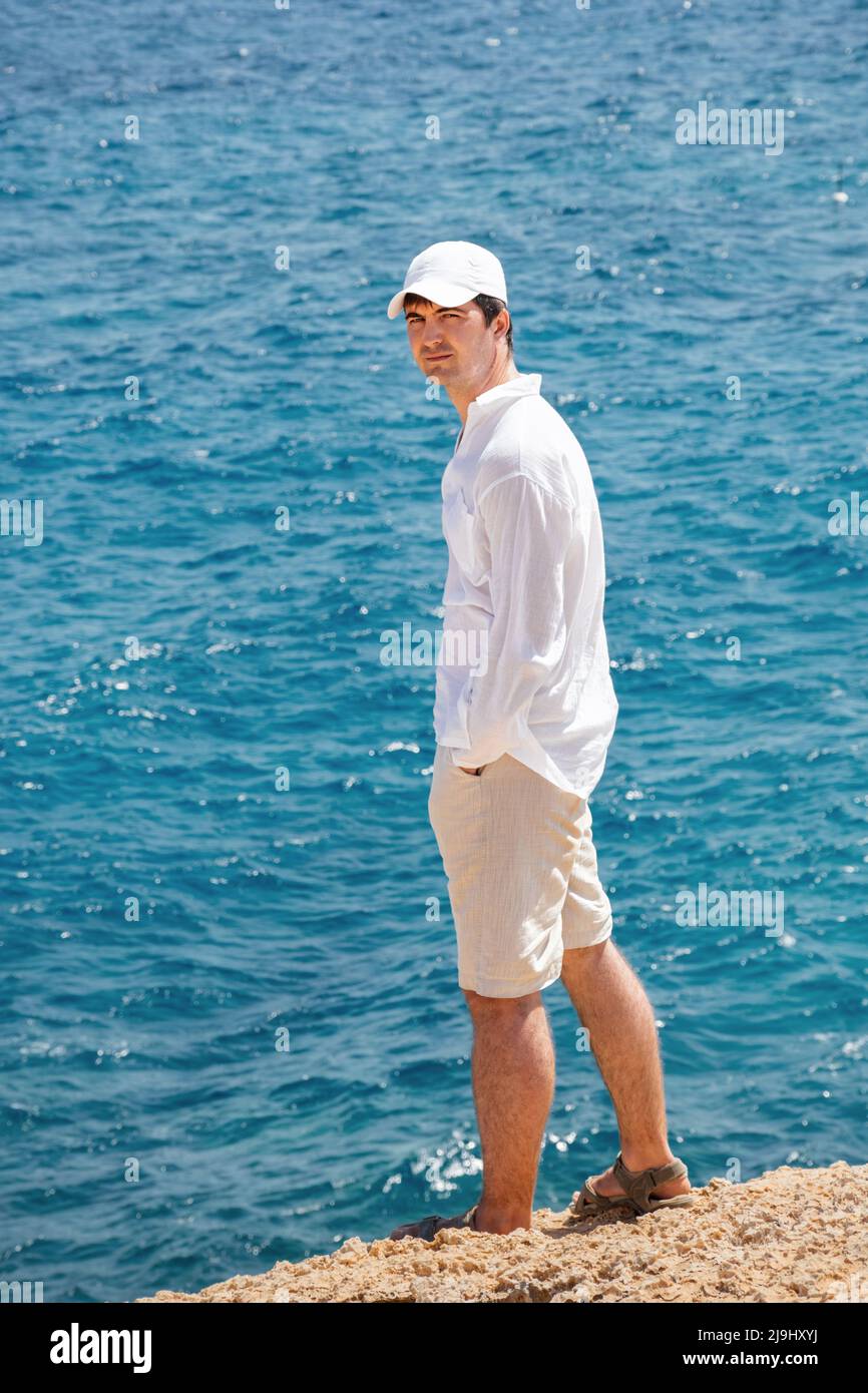 Vista posteriore di un uomo su uno sfondo di spiaggia rocciosa gialla e  mare blu. Un ragazzo in una camicia di lino bianco, pantaloncini, sandali e  un cappellino. Vacanze di viaggio. Mani