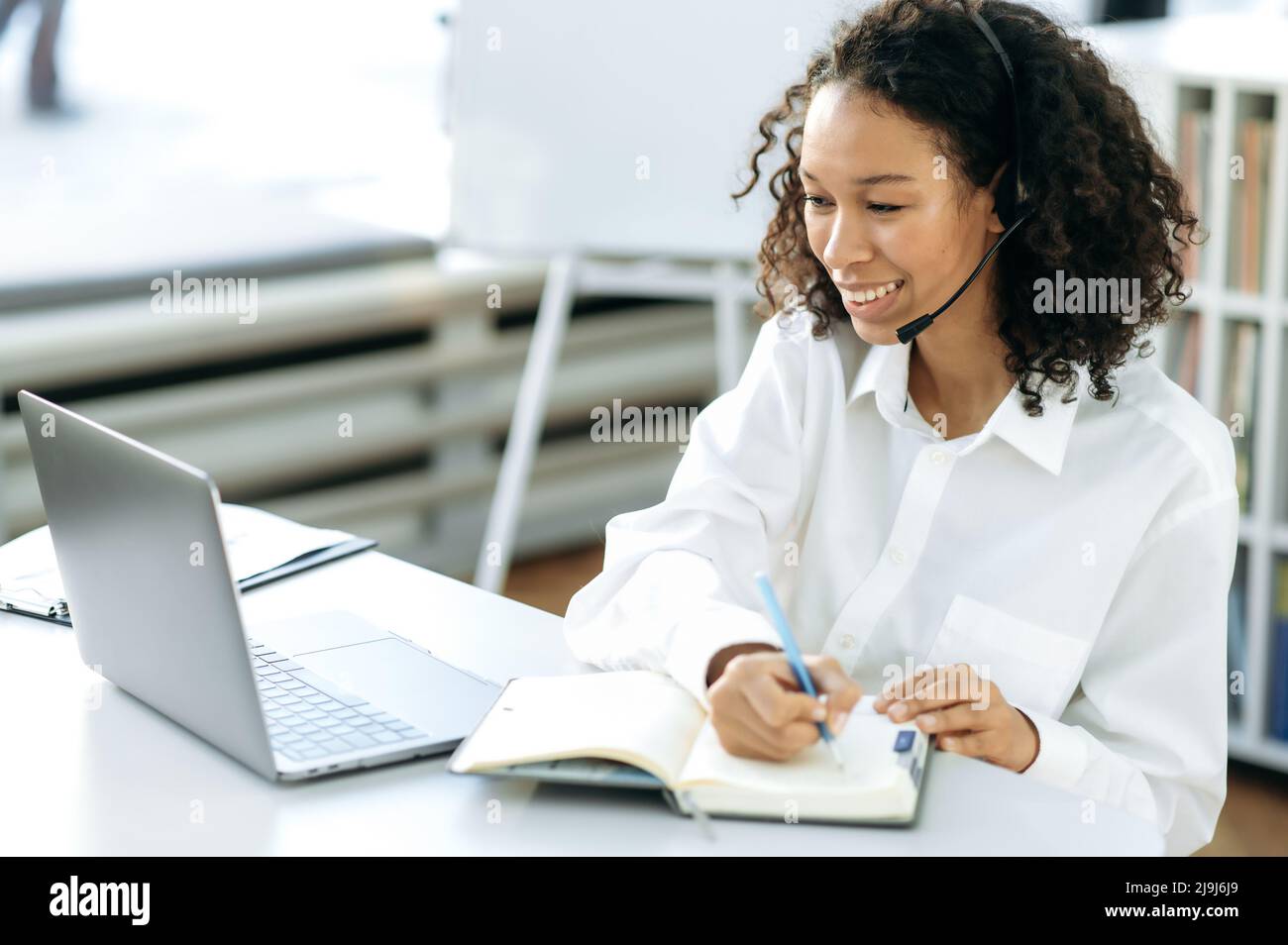Donna afro-americana amichevole positiva con capelli ricci, responsabile delle vendite online, addetto al servizio di assistenza, seduto a una scrivania di fronte a un computer portatile in un ufficio moderno, facendo appunti di lavoro Foto Stock