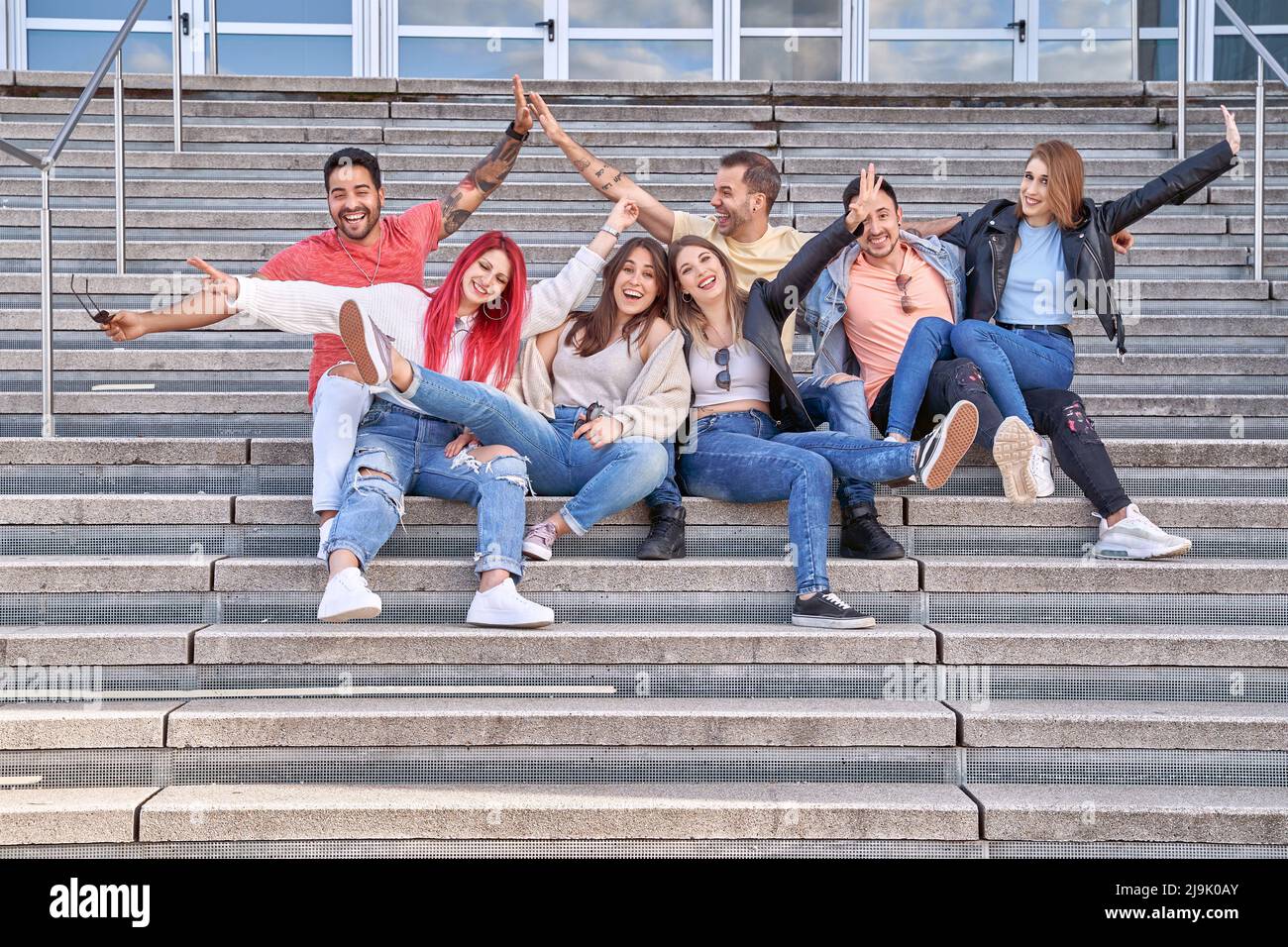 Gruppo di allegri giovani multi-etnici uomini e donne in abiti casual, sorridendo felicemente e guardando la macchina fotografica come si siedono sulle scale e si abbracciano dopo i loro studi di successo. Foto Stock