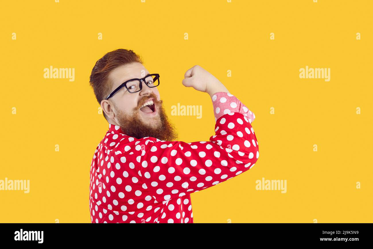Divertente ed elegante uomo barbato con sorriso sui visi che mostra bicipiti su sfondo giallo. Foto Stock