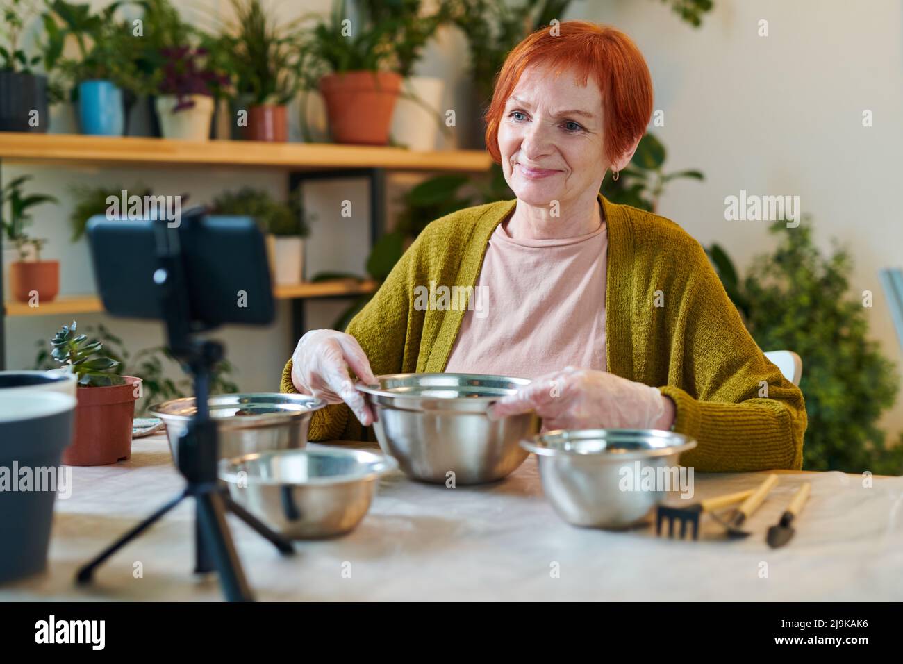 Redhead donna anziana seduta al tavolo con ciotole e video di tiro sul suo telefono cellulare, lei facendo blog sul giardinaggio Foto Stock