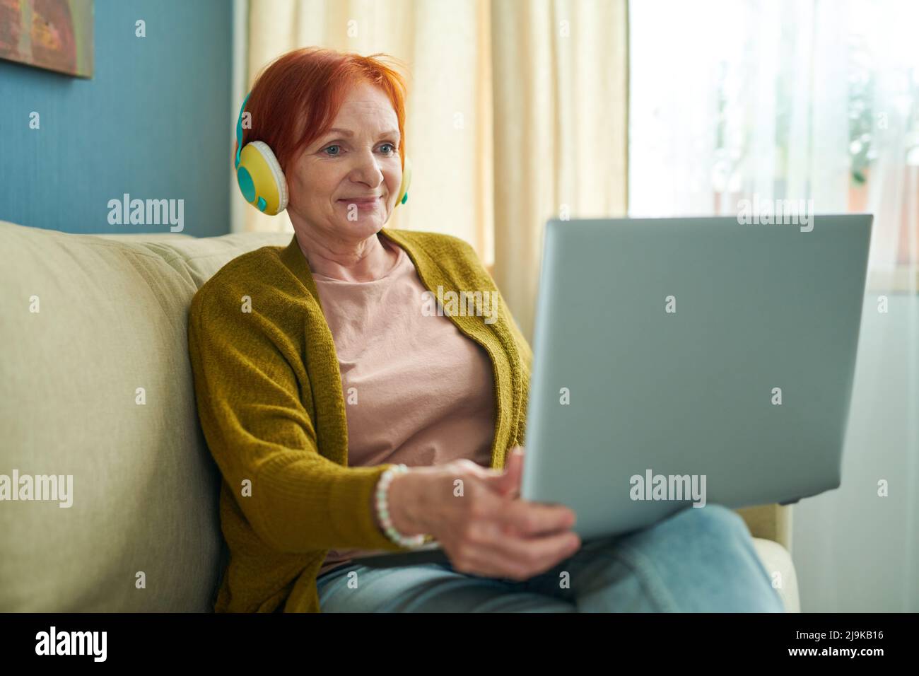 Donna anziana in cuffia seduta sul divano con computer portatile e parlare con i suoi parenti in linea durante l'isolamento Foto Stock