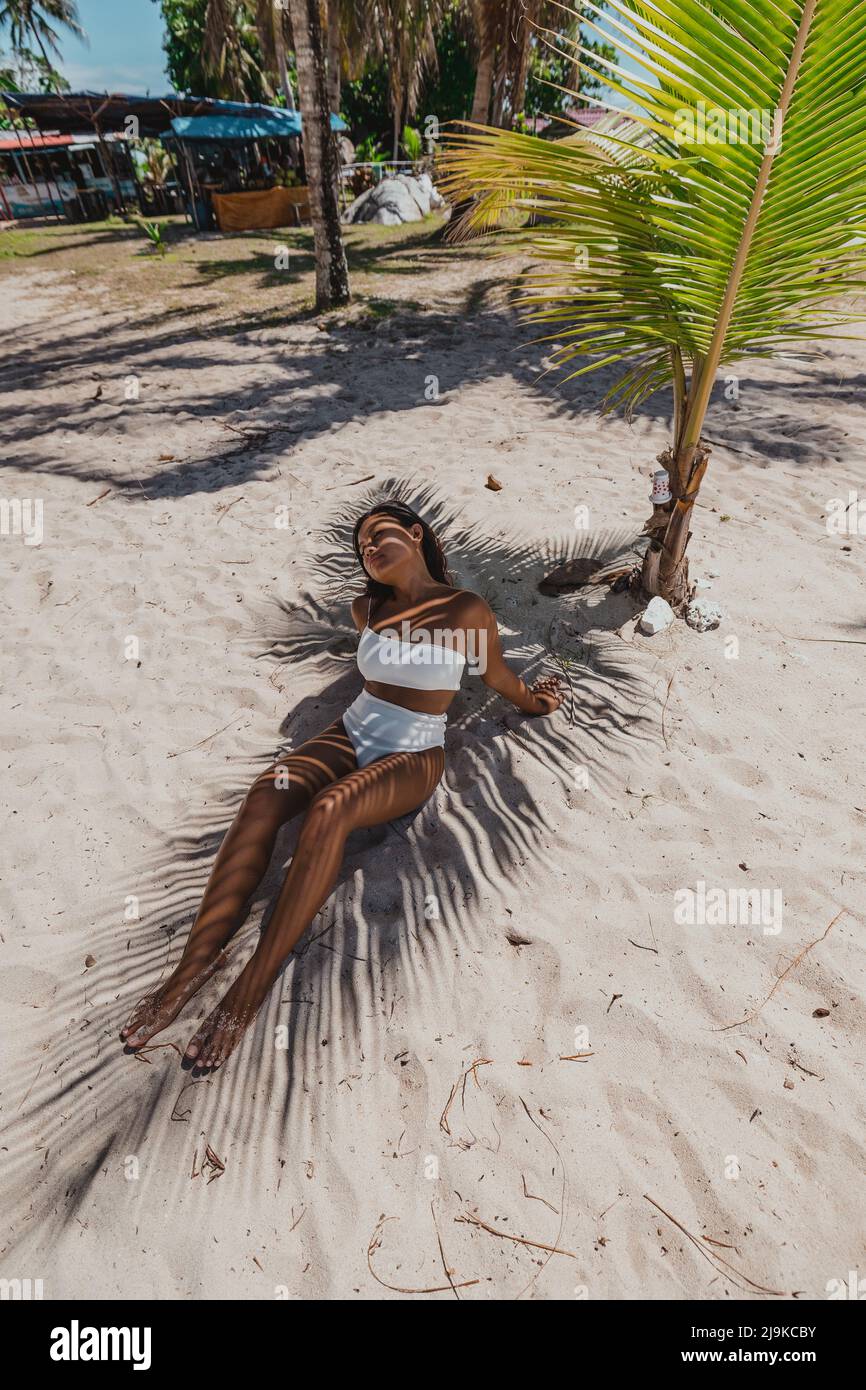 Giovane abbronzatura asiatica femmina sole bagnare in un bikini bianco sotto un ombra di palme su una spiaggia tropicale di sabbia bianca nell'Isola di Belitung Indonesia Foto Stock