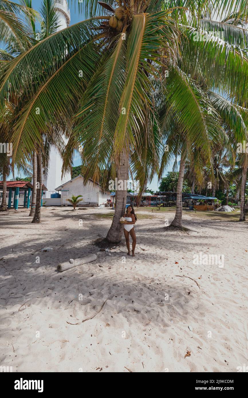 Giovane ragazza asiatica in piedi sotto un albero di cocco in un bikini bianco su una spiaggia di sabbia bianca tropicale a Belitung Island Indonesia Foto Stock