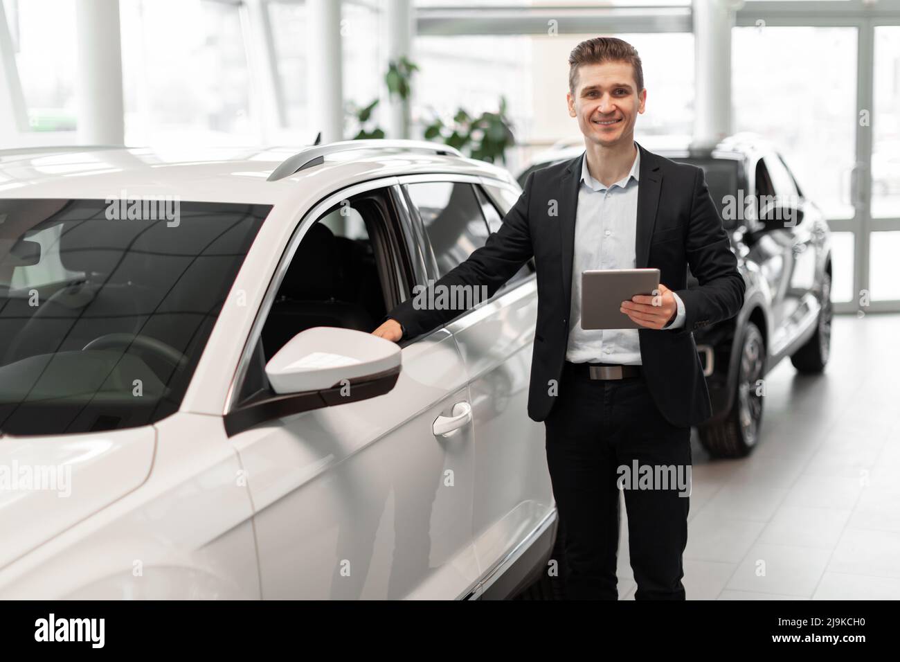 Allegro direttore delle vendite giovane con computer tablet in piedi vicino alla nuova automobile al salone di auto di lusso, spazio copia Foto Stock