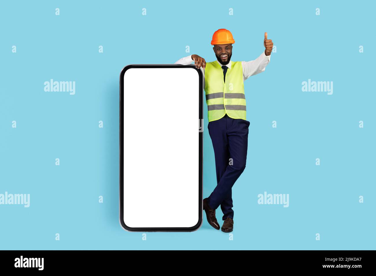 Sorridente ingegnere di costruzioni nero in piedi vicino a smartphone vuoto e mostrando Thumb Up Foto Stock