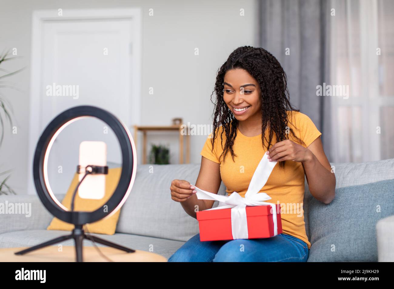 Allegra ragazza afroamericana famosa blogger che mostra ai seguaci i suoi regali di compleanno, utilizzando il telefono set sulla lampada ad anello Foto Stock