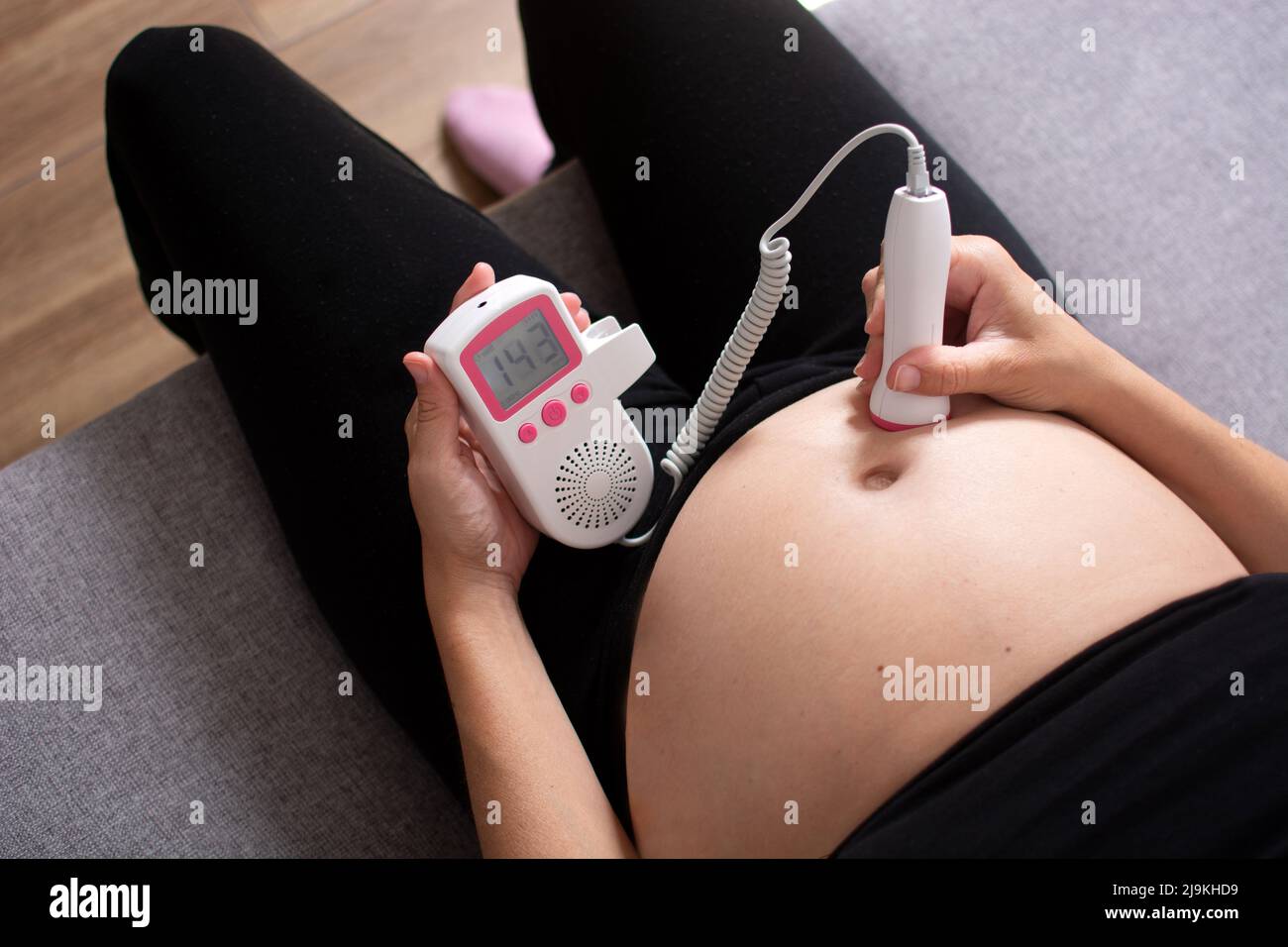 Donna incinta con doppler fetale che ascolta il cuore del bambino seduto sul divano a casa Foto Stock