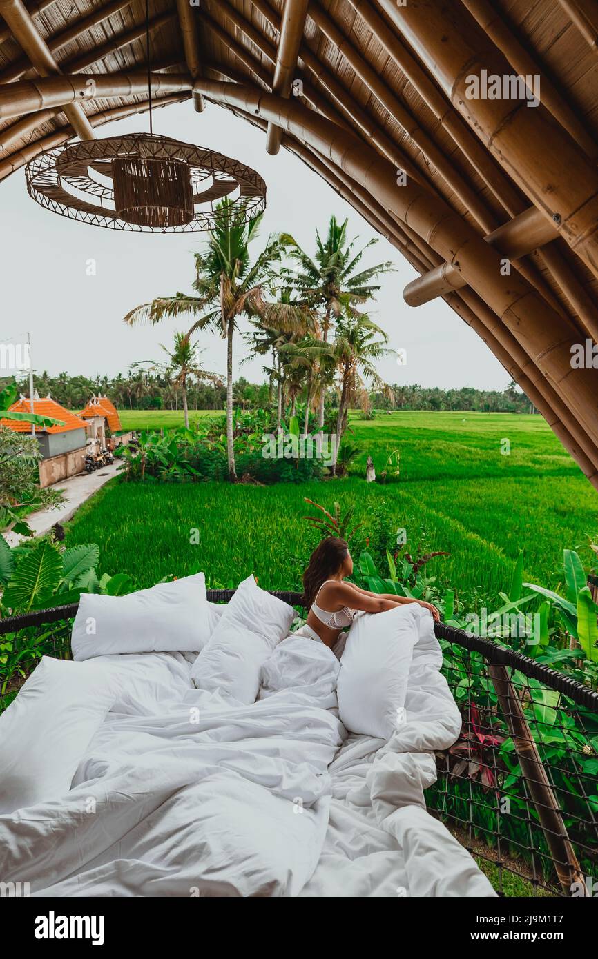 giovane donna asiatica con lenzuola bianche che si affacciano sui campi di riso di bali e sugli alberi di cocco dal suo balcone villa di bambù Foto Stock