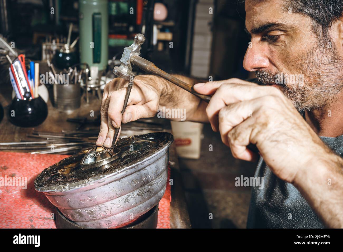 Raccolto uomo maturo bearded con martello e punzone per carve ornamento su argento dettaglio durante il lavoro in officina professionale Foto Stock