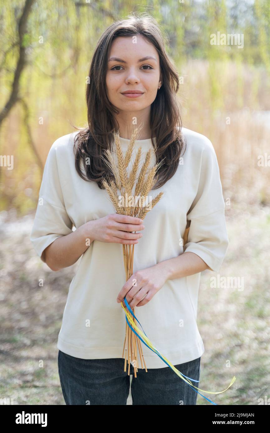 Giovane ragazza allegra con lunghi capelli scuri in abiti casual sorridente e guardando la macchina fotografica mentre in piedi in natura con un mazzo di spikelets di grano legato Foto Stock