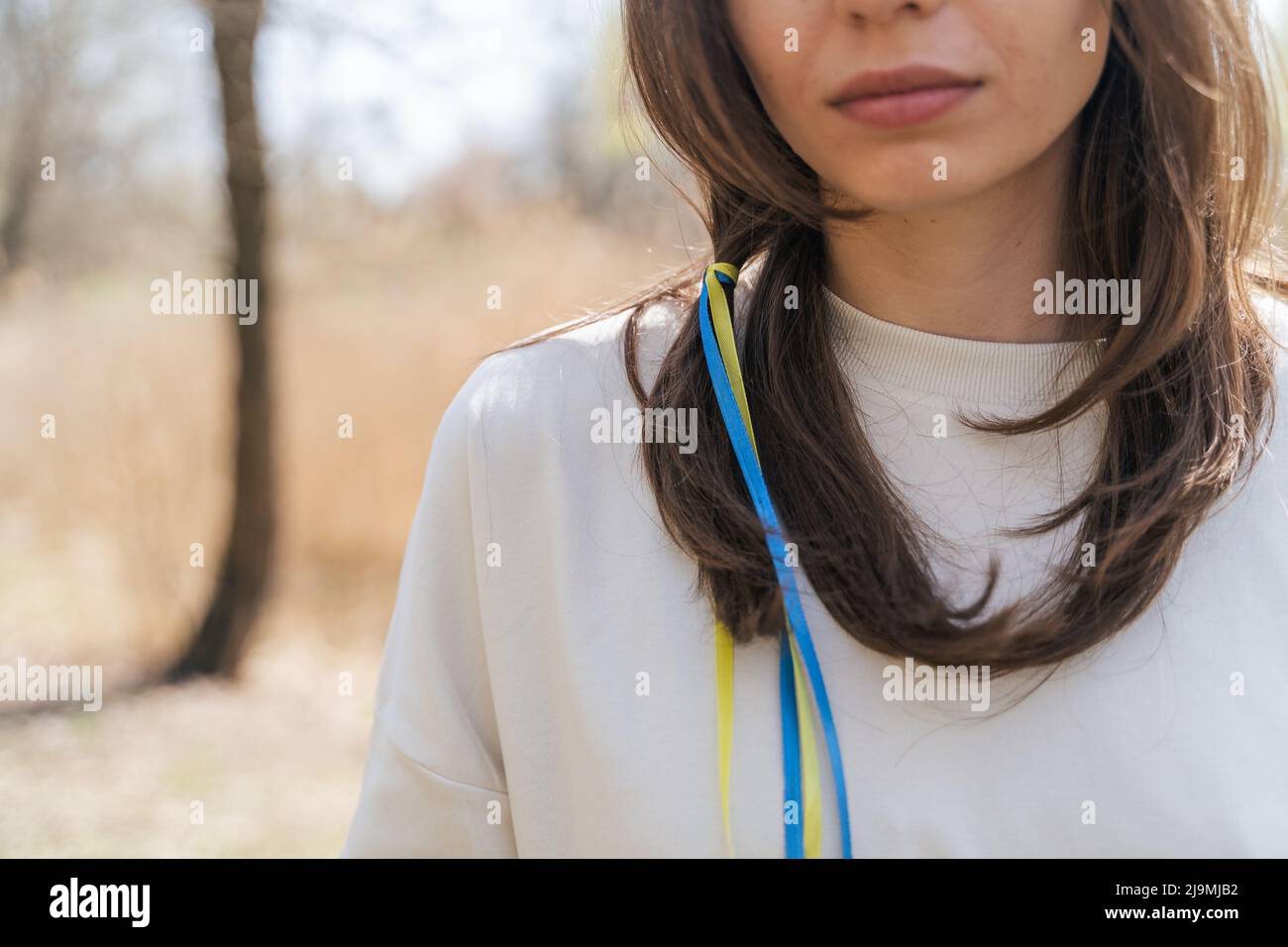 Calmo giovane donna in blusa bianca con nastri bandiera Ucraina in capelli lunghi marrone guardando la macchina fotografica mentre in piedi in campo in giorno di sole Foto Stock