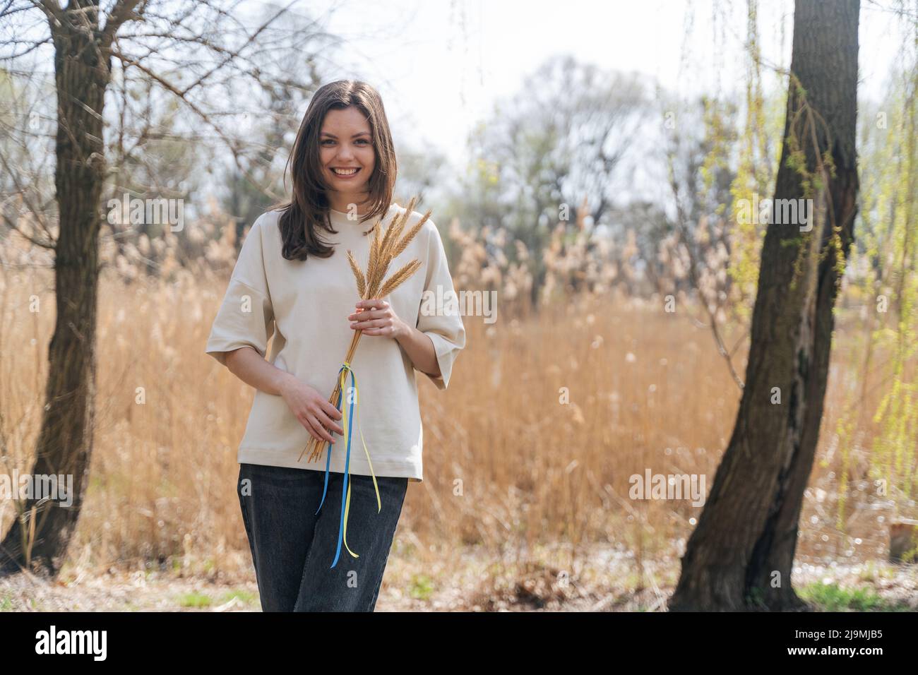 Giovane ragazza allegra con lunghi capelli scuri in abiti casual sorridente e guardando la macchina fotografica mentre in piedi in natura con un mazzo di spikelets di grano legato Foto Stock