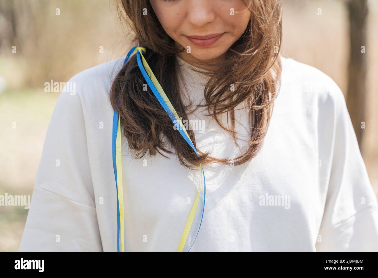 Calmo giovane donna in blusa bianca con nastri bandiera Ucraina in capelli lunghi e marroni sorridenti e guardando giù mentre in piedi in campo nella giornata di sole Foto Stock