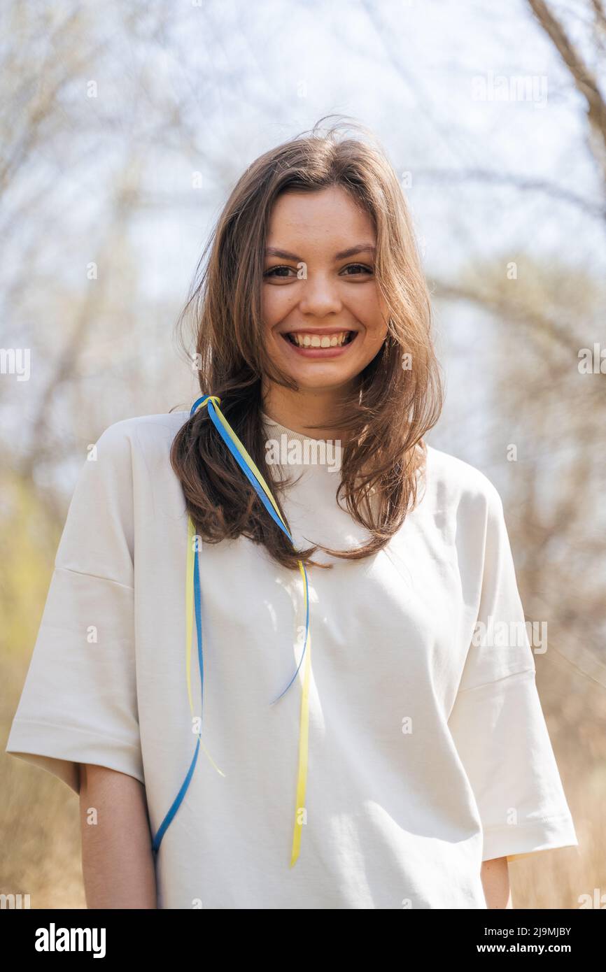Calma giovane donna in blusa bianca con nastri bandiera Ucraina in capelli lunghi marrone sorridente e guardando la macchina fotografica mentre in piedi in campo in giorno di sole Foto Stock
