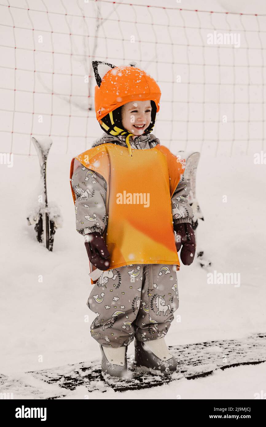 Corpo pieno di bambino allegro in caldo outerwear e arancione casco protettivo e gilet sorridente mentre si trova in piedi su terreno nevoso prima di sciare in winte Foto Stock