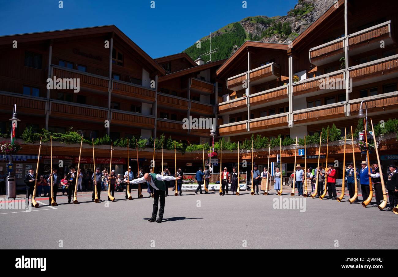 I musicisti svizzeri suonano l'alphorn, uno strumento musicale tradizionale guidato da un direttore del coro sulla strada principale del villaggio di Zermatt. Foto Stock