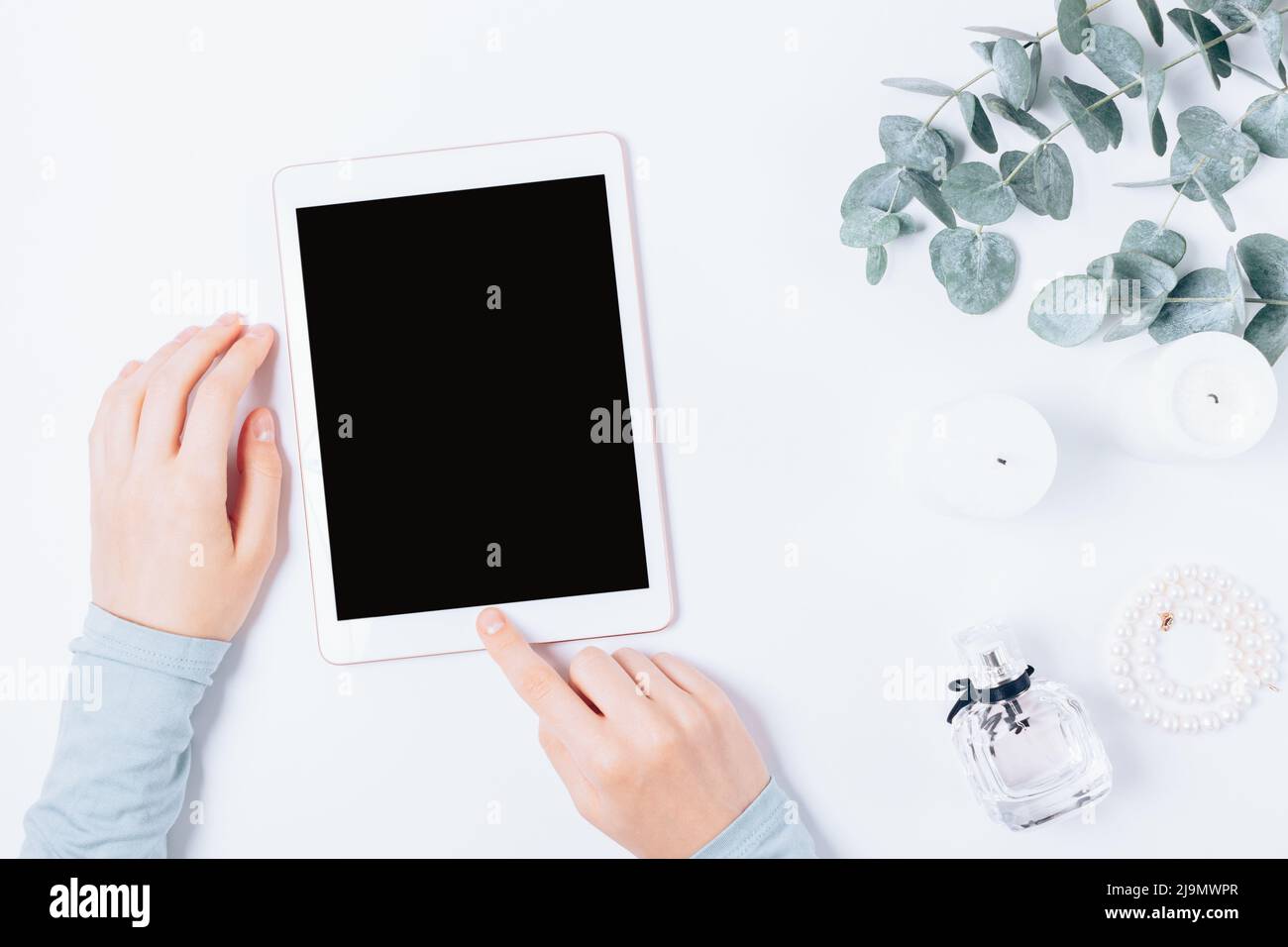 Le mani della donna tengono il tablet elettronico accanto agli accessori femminili e ai ramoscelli in eucalipto verde sul tavolo bianco, vista dall'alto. Foto Stock