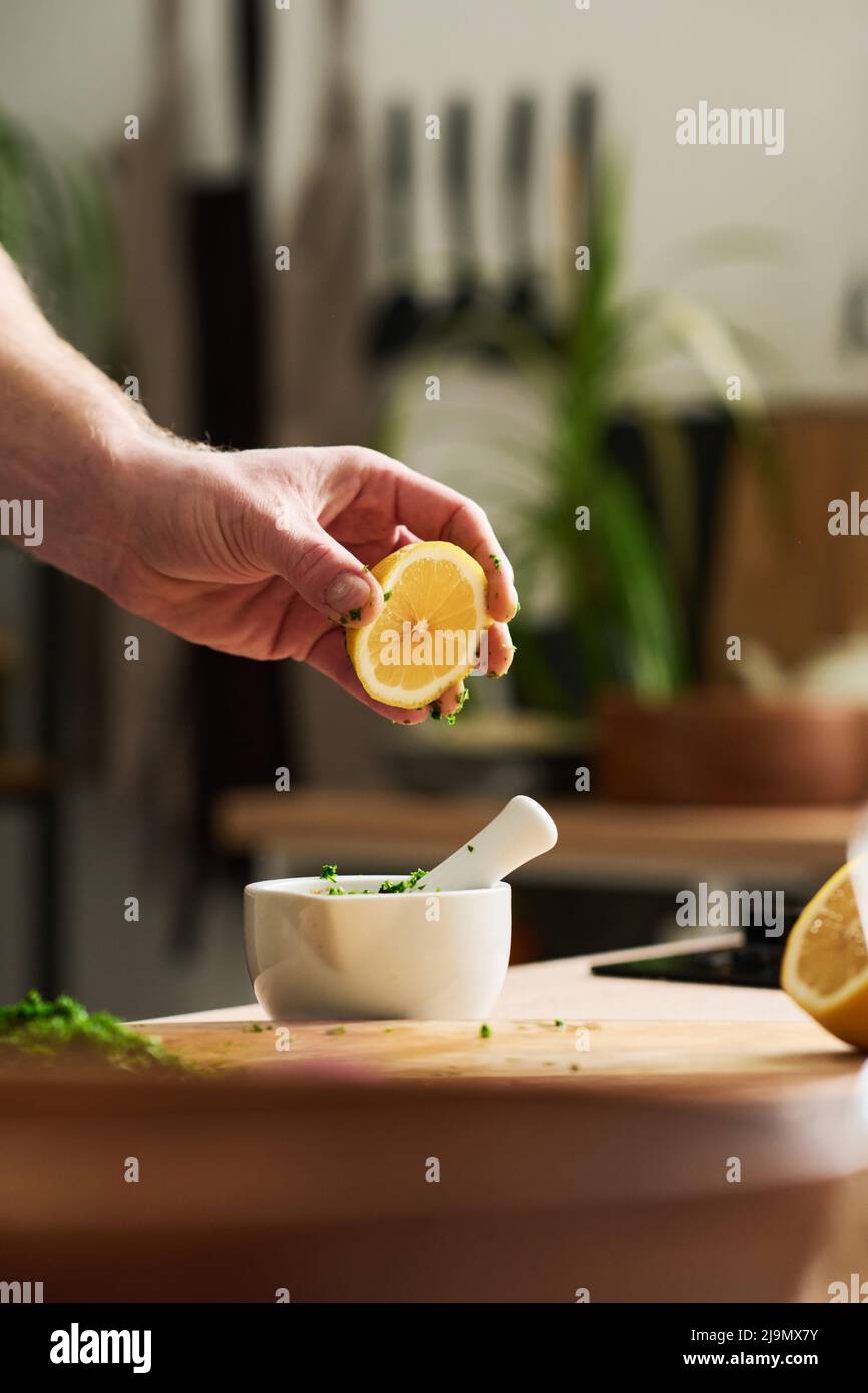 Primo piano verticale medio di un uomo irriconoscibile che tiene un pezzo di limone sopra il mortaio con spezie e coriandolo tritato per spremere il succo Foto Stock