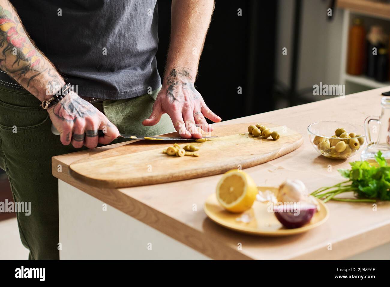 Uomo tatuato irriconoscibile che taglia olive sottaceto su tavola di legno in cucina mansarda, a sezione media Foto Stock