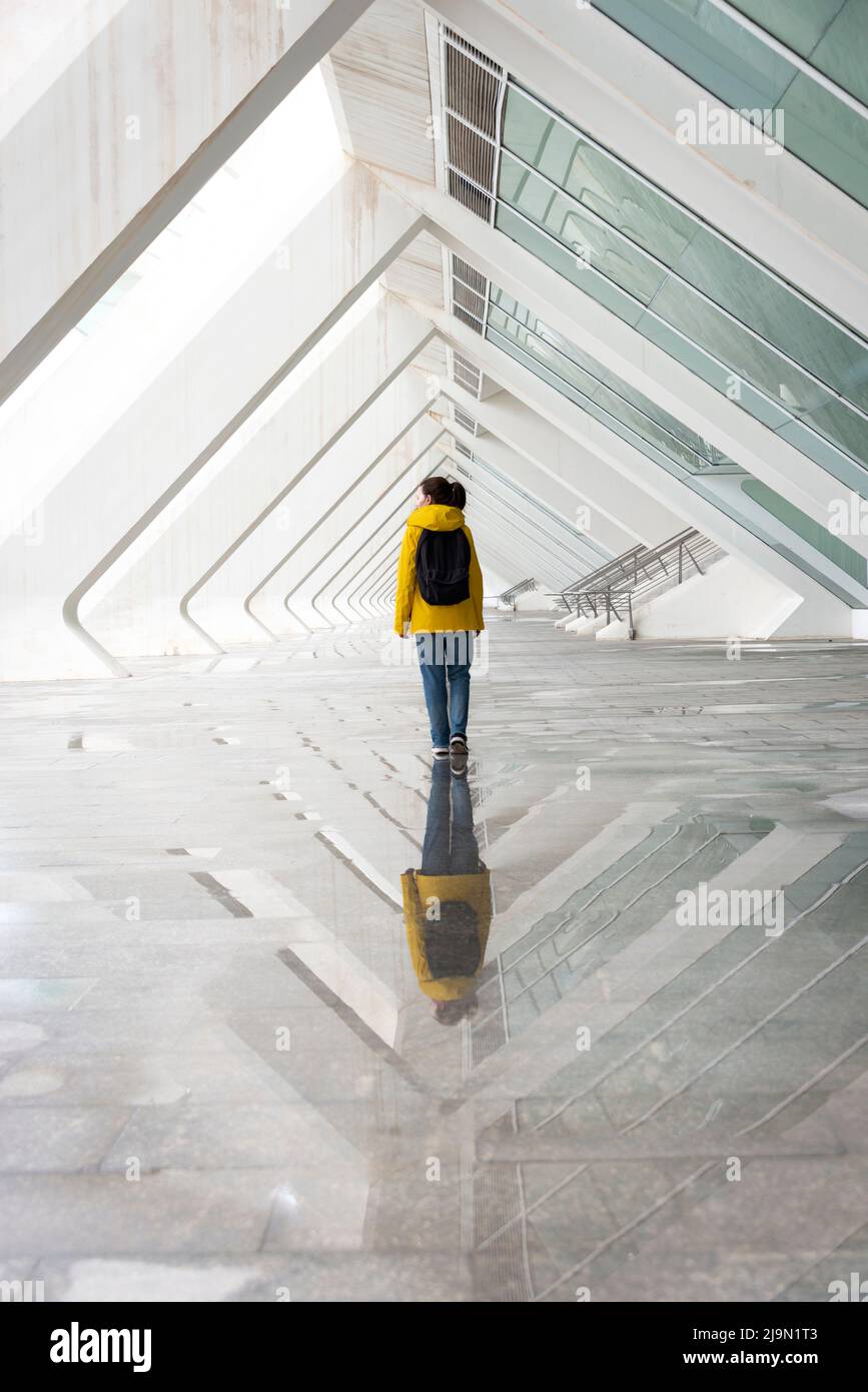 vista posteriore di una donna che cammina attraverso l'architettura moderna sotto la pioggia indossando un cappotto giallo. Foto Stock