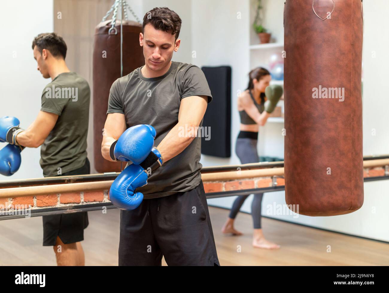 Determinato atletico maschio mettendo su guanti di boxe mentre si prepara per l'allenamento e in piedi vicino sacco di punzonatura in palestra Foto Stock