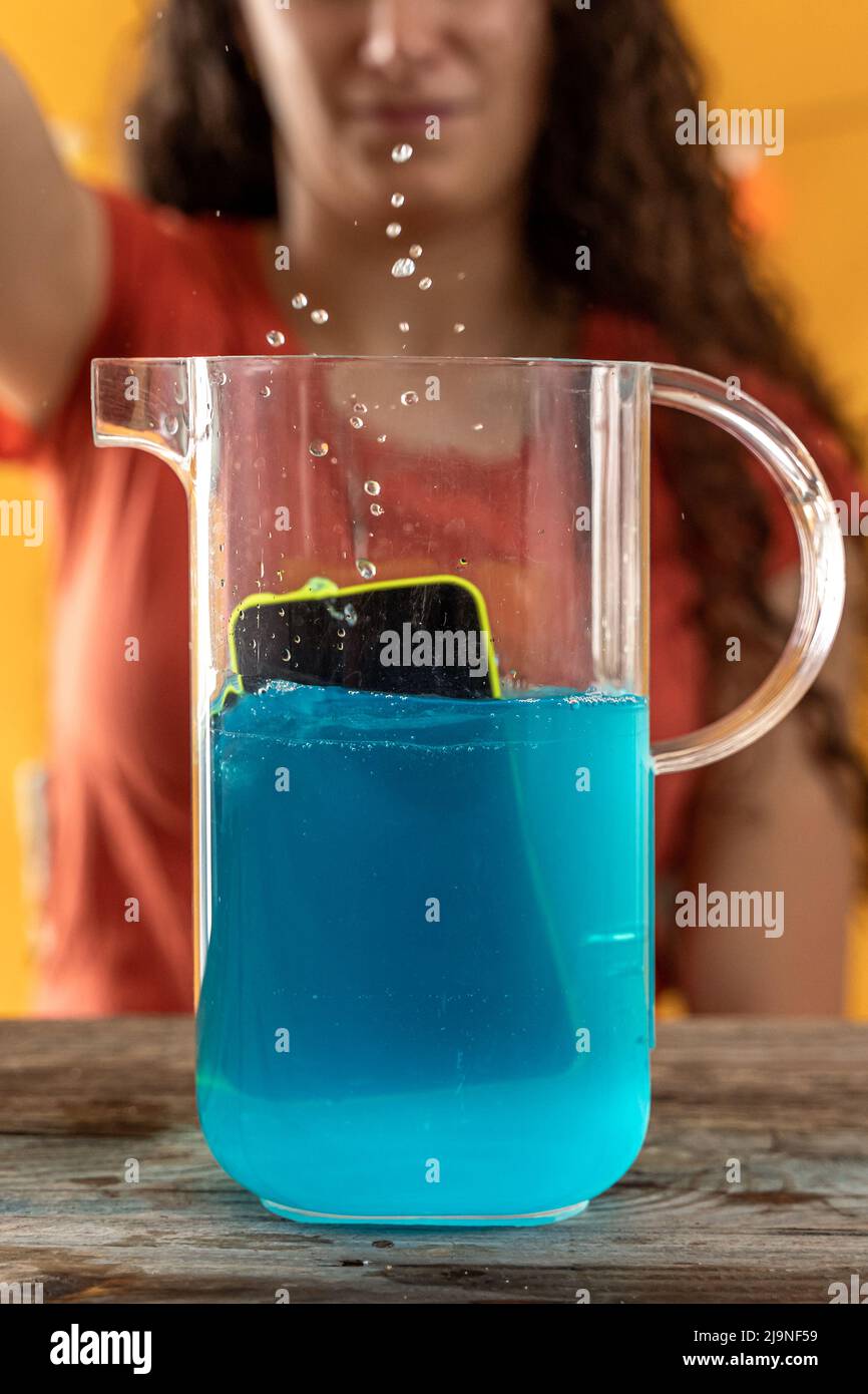 la giovane donna getta uno smartphone in una caraffa piena di un cocktail blu - scatto da studio Foto Stock