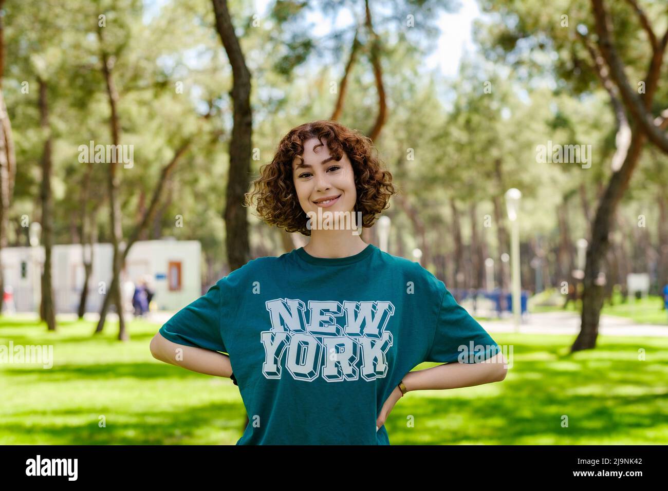 Ritratto di giovane donna rossa felice indossando una t-shirt verde in piedi sul parco della città, tenendo le mani all'aperto sui fianchi e guardando la macchina fotografica con sorrisi grandi Foto Stock
