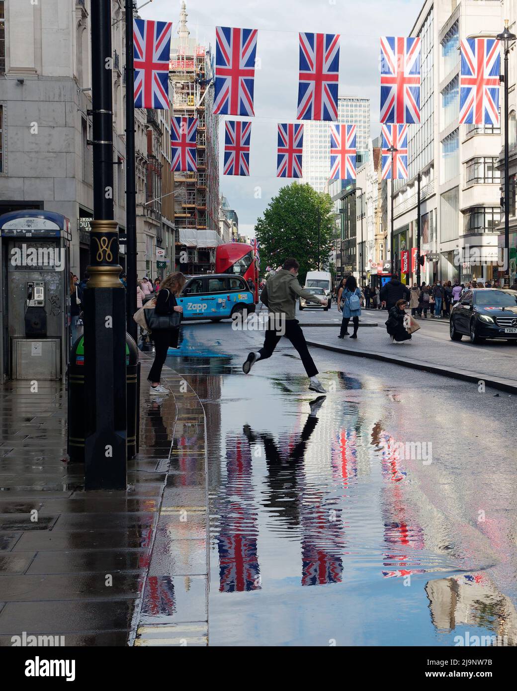 Mans salta attraverso le pozze d'acqua a Oxford Street dopo una pioggia pesante, con le bandiere di Union Jack riflesse nell'acqua. Foto Stock