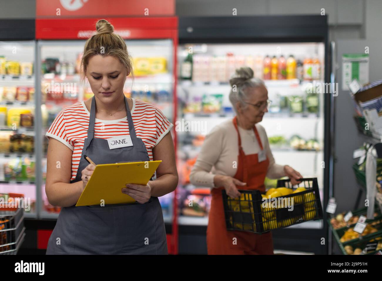 Giovane assistente di negozio in supermercato in guscio di verdure, in pankground è il suo collega riempimento scorte. Foto Stock