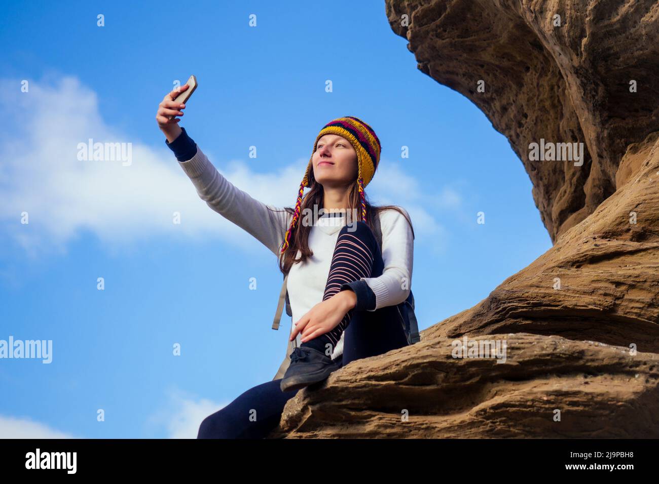 Bella sottile e sportiva giovane donna turistica in un divertente cappello da Nepal lana yak seduta e hanno riposo facendo selfie telefono arrampicata grande roccia su Foto Stock