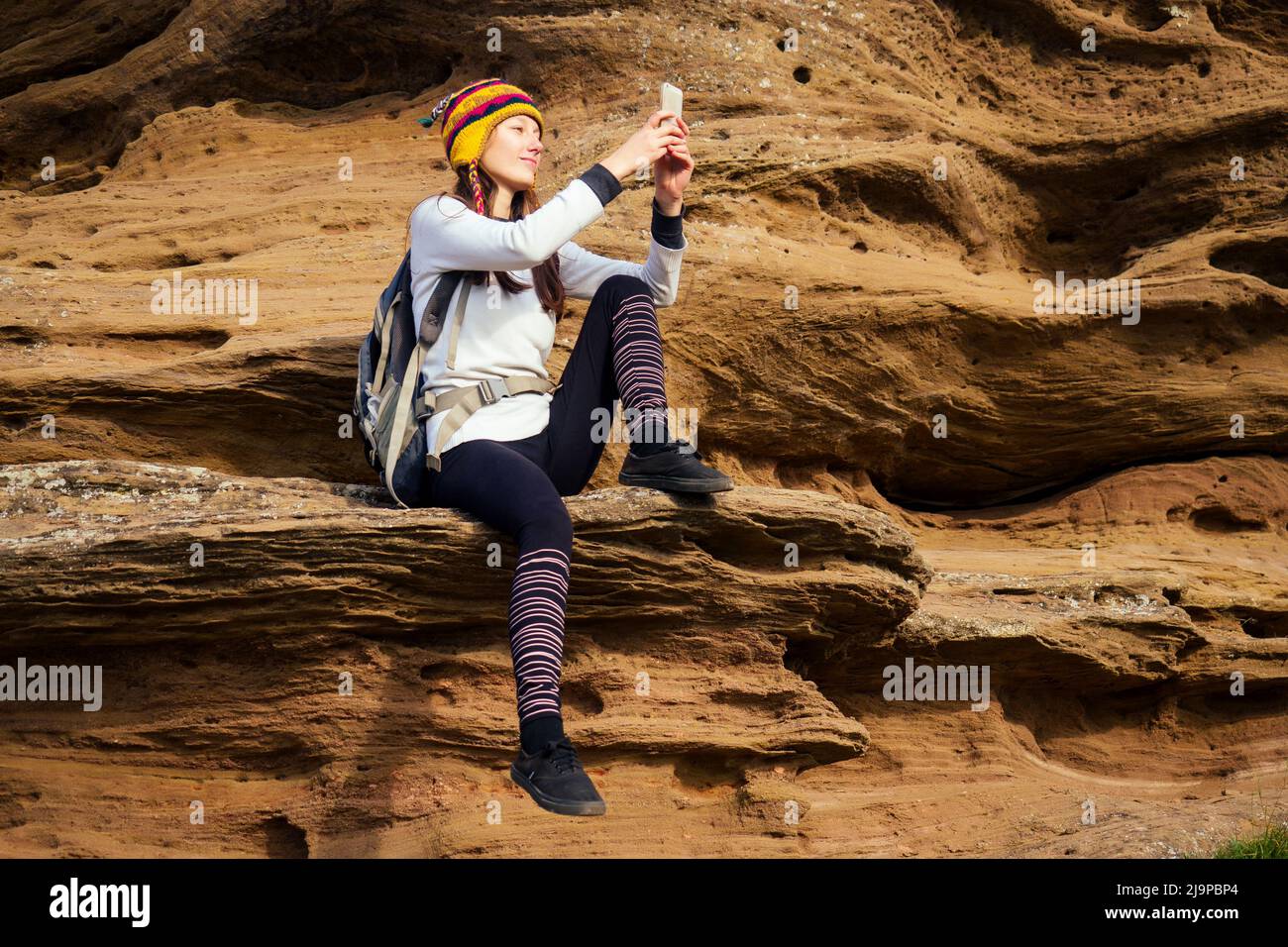 Bella sottile e sportiva giovane donna turistica in un divertente cappello da Nepal lana yak seduta e hanno riposo facendo selfie telefono arrampicata grande roccia su Foto Stock