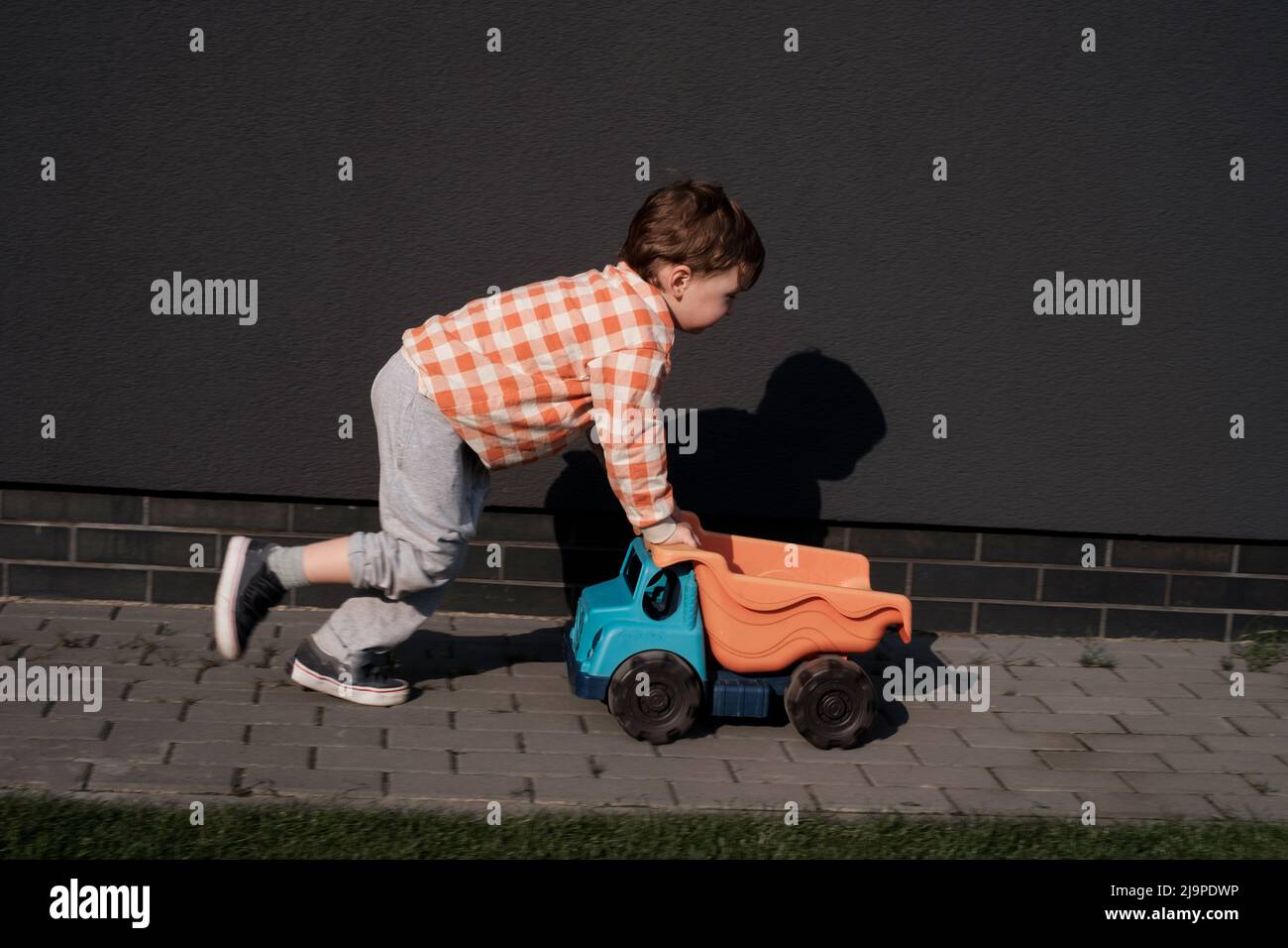 Ragazzo che gioca con l'auto giocattolo vicino a casa sopra il muro nero. Bambini piccoli all'aperto divertirsi. I bambini trascorrono del tempo sull'aria fresca nell'area urbana. Giorno di sole luminoso Foto Stock