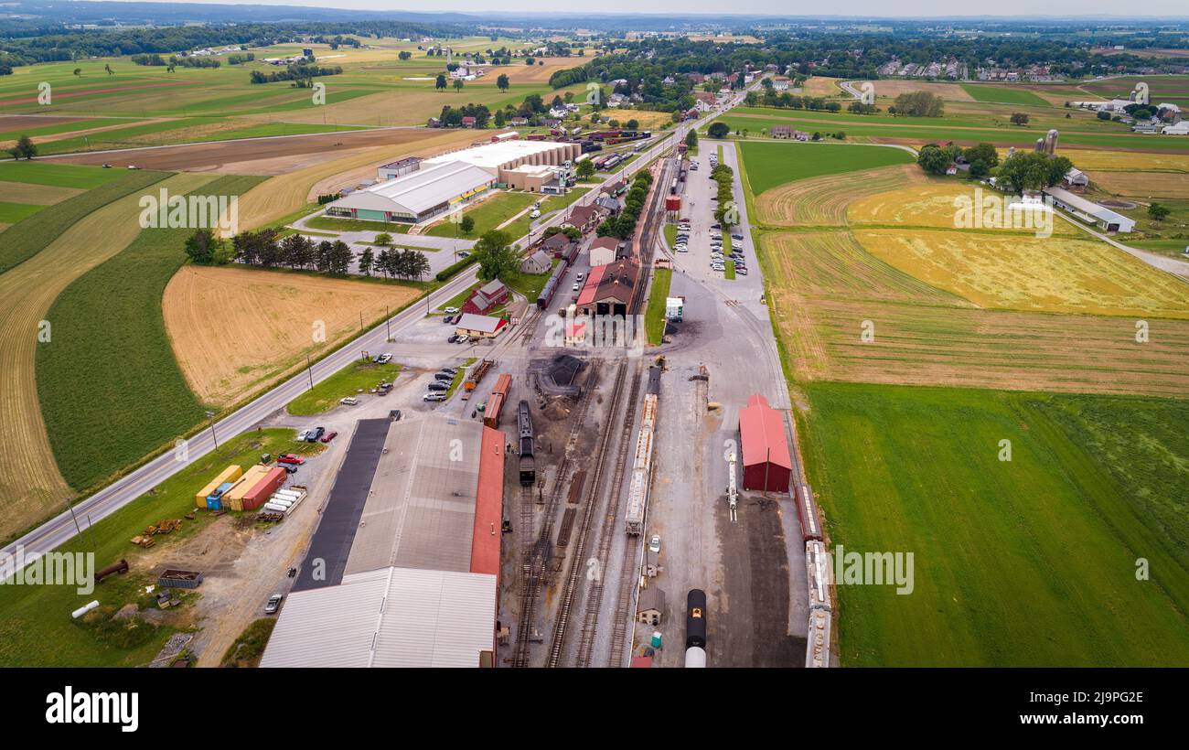 Vista con droni di una stazione passeggeri a vapore e di un campo merci con materiale rotabile e piste vuote in un giorno estivo nuvoloso Foto Stock