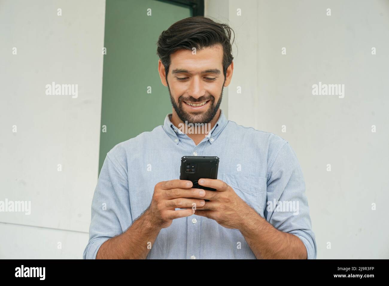 Uomo d'affari sorridente con smartphone. Bel dirigente che guarda lontano mentre si alza contro il muro. Indossa astuti casuals. Foto Stock