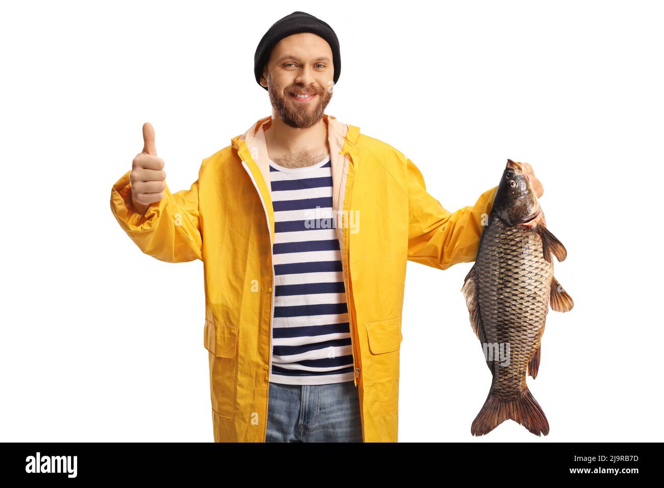 Giovane pescatore in un impermeabile giallo che tiene un pesce di carpa e che gestura i pollici su isolato su sfondo bianco Foto Stock