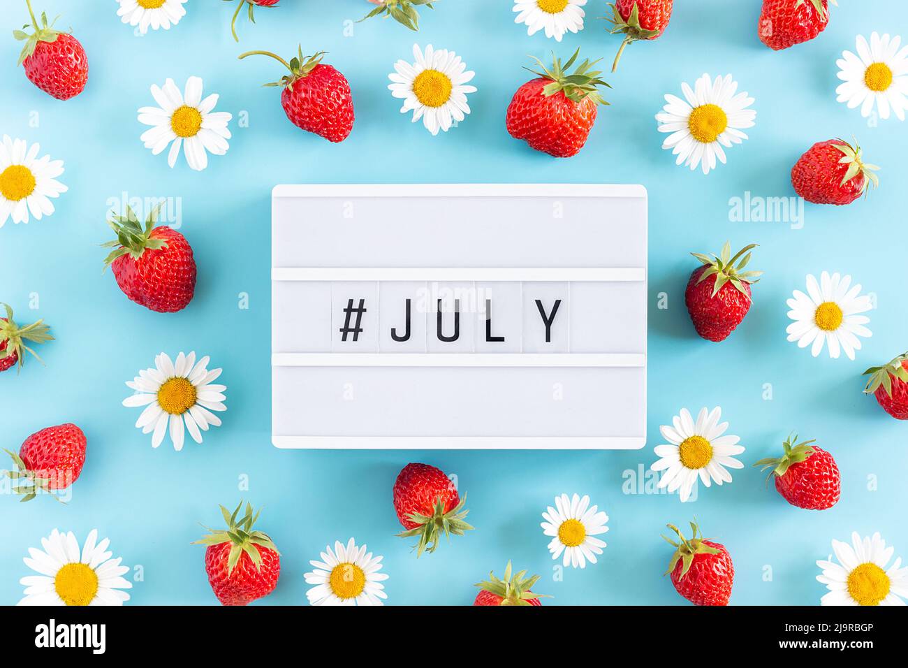 Estate mese Luglio testo su scatola leggera e fragola, fiori di camomilla su sfondo blu. Concetto creativo Ciao luglio. Vista dall'alto, piatto, saluto ca Foto Stock