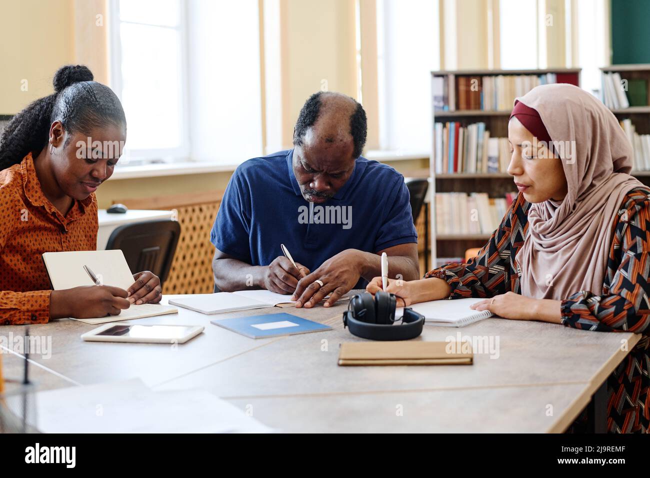 Gruppo di tre studenti immigrati multietnici seduti a tavola in biblioteca che svolgono un compito di scrittura durante la lezione Foto Stock