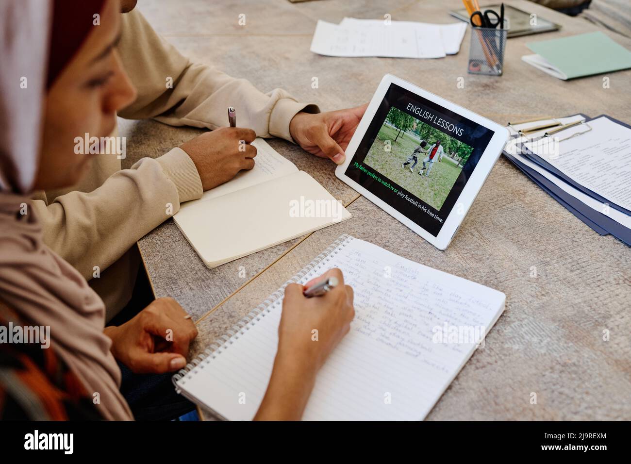Donna musulmana e il suo compagno di classe irriconoscibile che guarda il video educativo su tablet digitale e che prende appunti durante la lezione di inglese Foto Stock