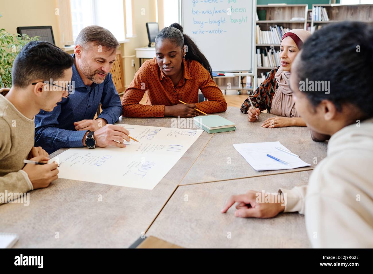 Insegnante di lingua inglese seduto al tavolo con i suoi studenti che controllano l'ortografia delle parole sul poster che hanno creato Foto Stock