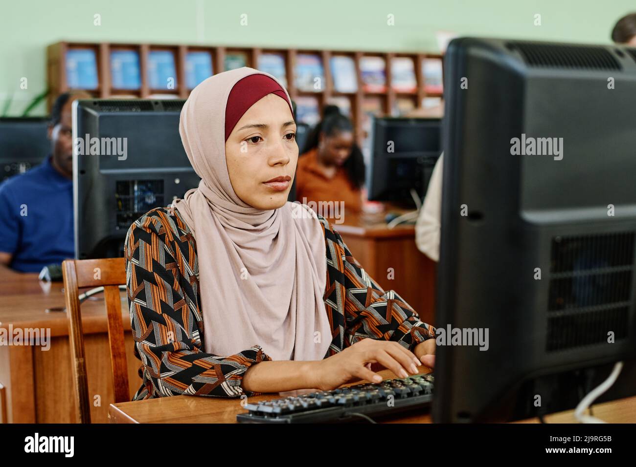 Ritratto di donna musulmana pensiva che indossa hijab seduto in biblioteca universitaria testando qualcosa sul computer desktop Foto Stock