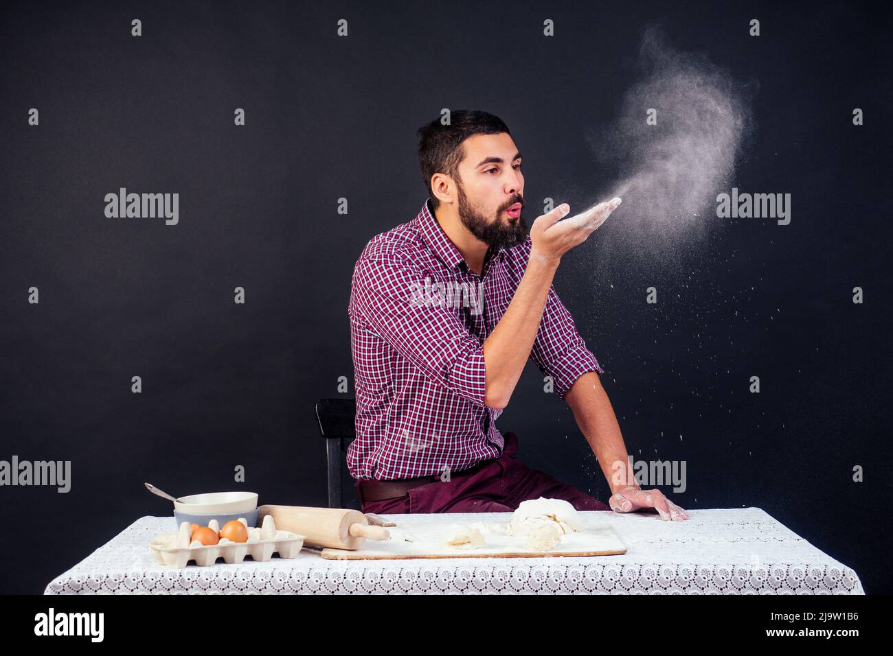 giovane e attraente panettiere caucasico con barba fa l'impasto cosparso di farina su sfondo nero in studio. La pasticceria cuoce la torta Foto Stock